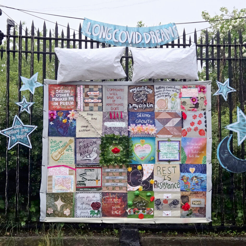 Photo of a metal fence in front of trees and bushes, with an art installation hanging on it. The installation has a handpainted blue banner across the top with white text that says: "LONG COVID DREAMS." Below this is artwork representing a bed - there are two white pillows and a colorful patchwork quilt made of small artworks. Surrounding the quilt are handpainted moons and stars.