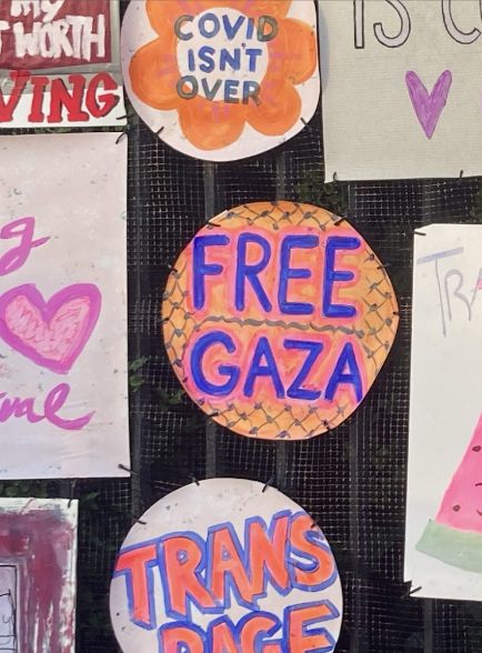 Image description: Photograph of “Free Gaza” sign before it was removed from the cemetery fence. The handpainted sign is a circular shape with an orange background with the black ‘fishnet’ pattern from a keffiyeh. Bold blue text outlined with pink says: “Free Gaza.”