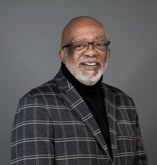 Photo of Sylvester, a Black person with a bald head and salt-and-pepper beard, wearing glasses and smiling. Sylvester wears a black sweater with a charcoal gray windowpane plaid blazer.