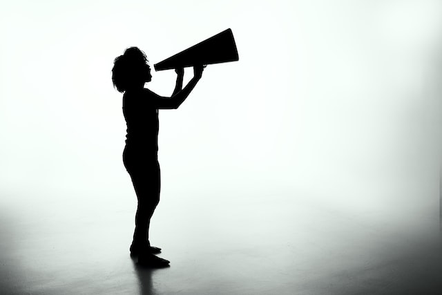 A black silhouette of a person speaking into a megaphone against a hazy grey background. 